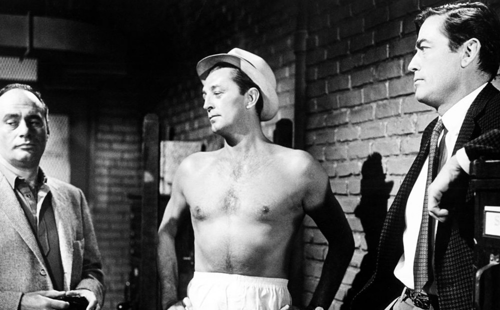 A shirtless Robert Mitchum stands inside with Gregory Peck and another man.