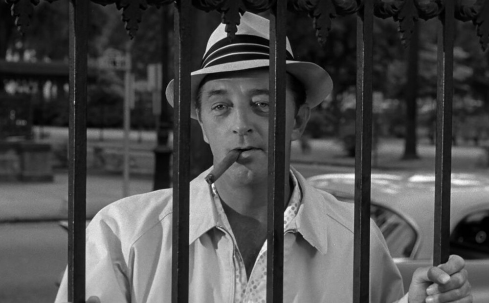 Actor Robert Mitchum, dressed all in white, smokes a cigar and holds onto the bars of a gate outside.