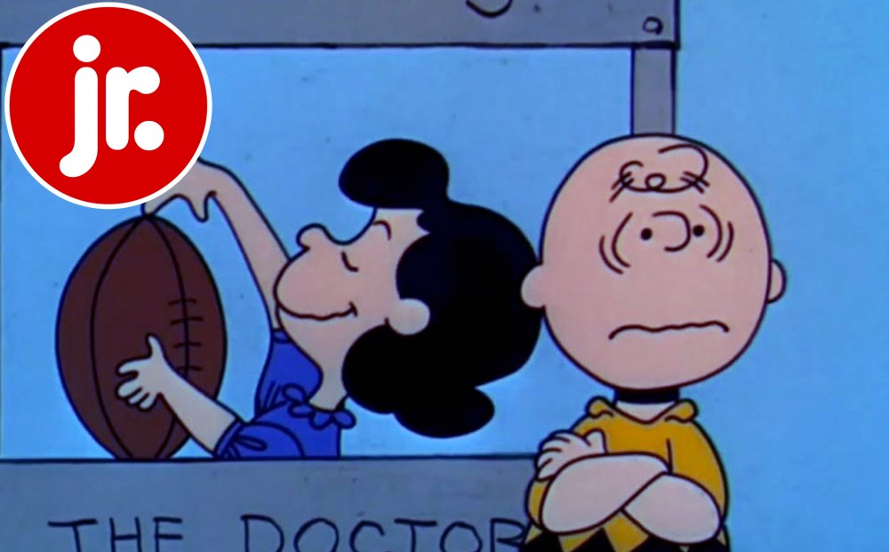 Lucy spins a football; Charlie Brown stands near her with his arms crossed.