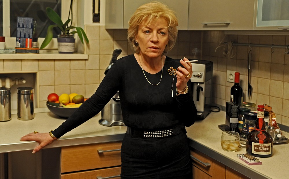 A woman in a black dress stands in a kitchen smoking a cigarette, with a tumbler of Grand Marnier on the counter next to her.