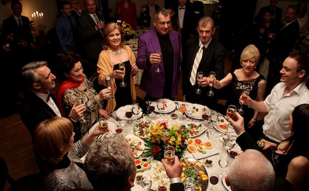 A group of people in dressy clothing stand around a table of hors d'ouevres raise glasses of champagne for a toast.