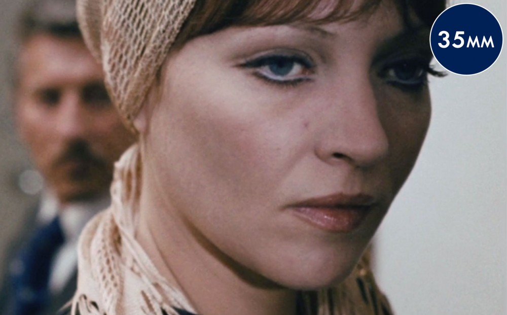 Close up on actor Anna Karina's face; a man appears behind her, out of focus.