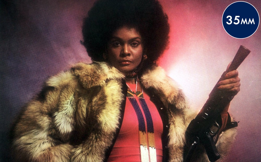 Actor Tamara Dobson stands imperiously, wearing a fur coat and holding a rifle under her shoulder.