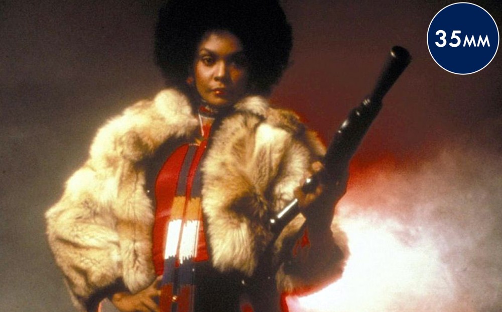 Actor Tamara Dobson wears a fur coat and holds a rifle.
