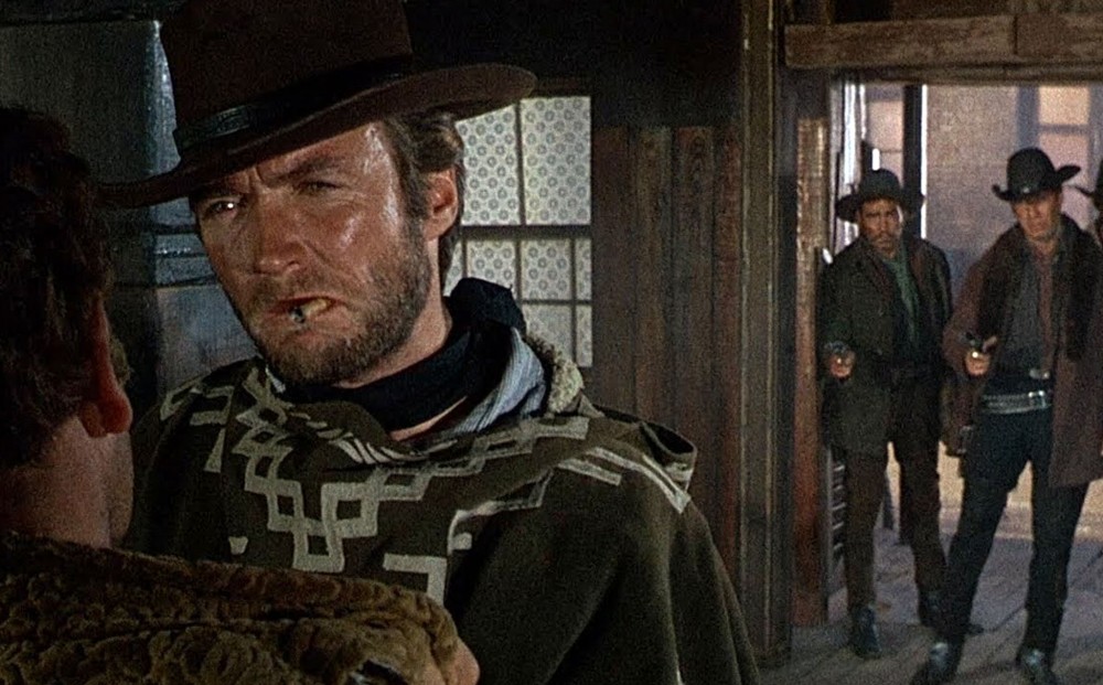 Actor Clint Eastwood speaks to someone in a saloon, his back to two men entering with their guns drawn and pointed at him.