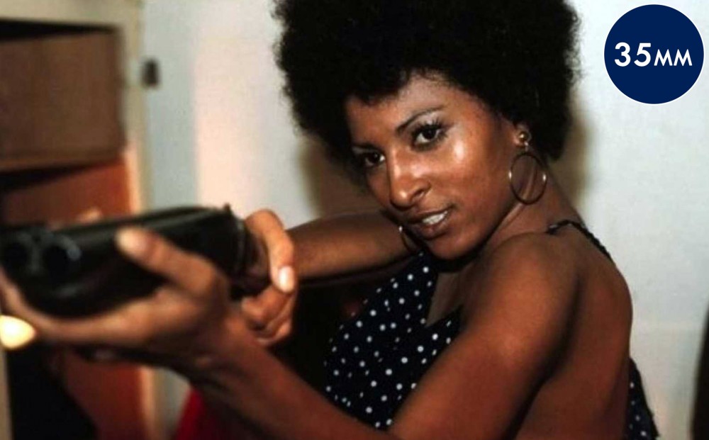 Actor Pam Grier aims a double-barreled gun at something off-camera.