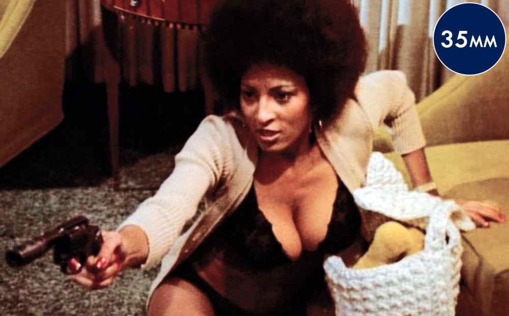 Actor Pam Grier sits on a living room floor, as though she has fallen, but aims a handgun at someone off-camera.