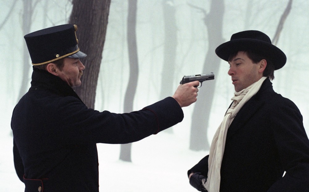 Actor Klaus Maria Brandauer points a gun at another man; they stand in a snowy forested area.