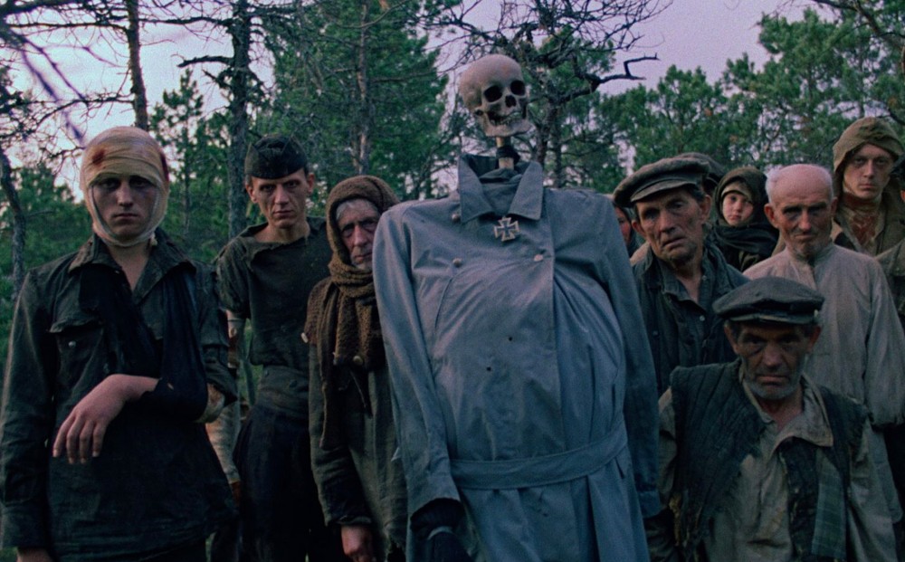A group of grizzled-looking citizens, some injured, stand in a clump; they hold up a sort of mannequin with a human skull as its head.