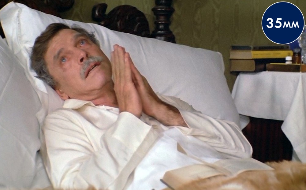 Actor Burt Lancaster lays on a bed with white sheets, wearing white pajamas; he holds his hands close to his face in a prayer position.