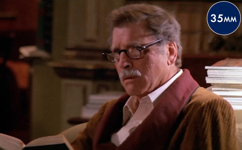 Actor Burt Lancaster sits in a study, wearing glasses and holding a book.