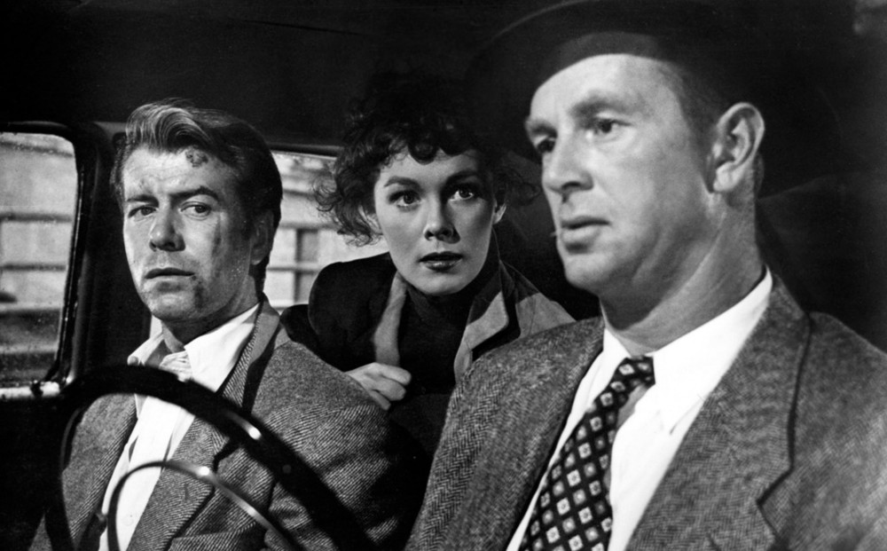 Two men sit in the front seat of a car, and a woman sits in the back seat, looking at the driver.