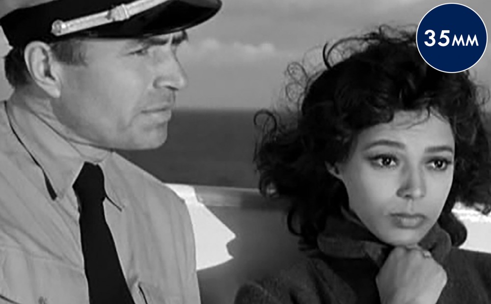 Actors Dorothy Dandridge and James Mason stand by the sea; she clutches her collar around her neck, and he wears a captain's uniform.