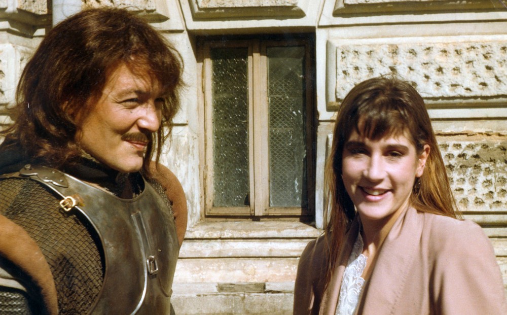 A man and woman stand next to each other, smiling; she is wearing regular clothes and he appears to be wearing a knight's costume.