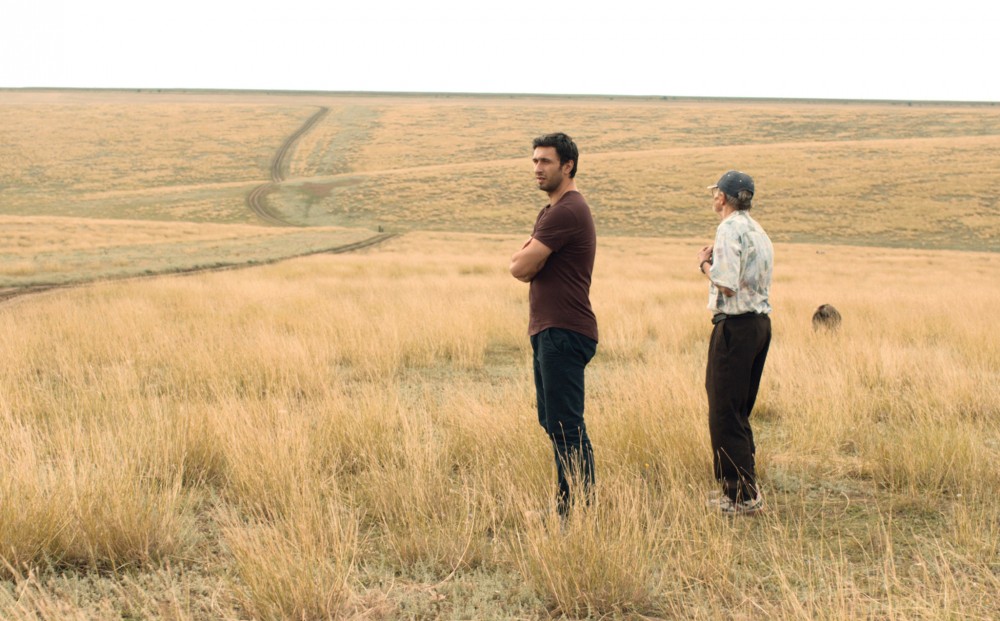 A middle-aged man and an older man stand int he middle of a vast, empty field.
