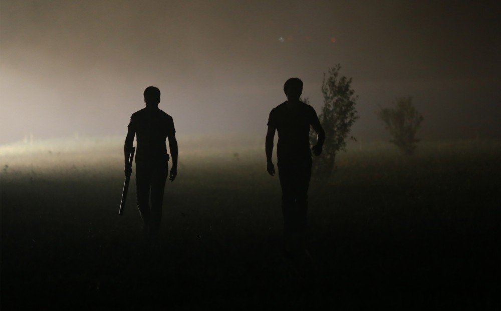 At night, two men appear as dark shadows, walking through a field.