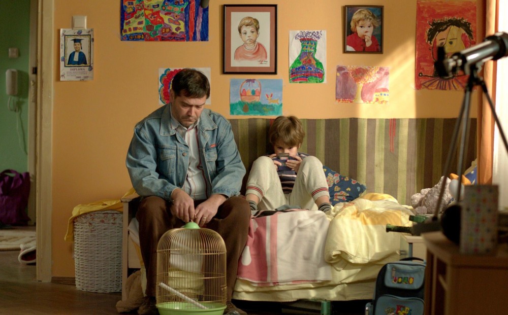 A man and a child sit in a colorful child's bedroom; the man holds an empty birdcage.