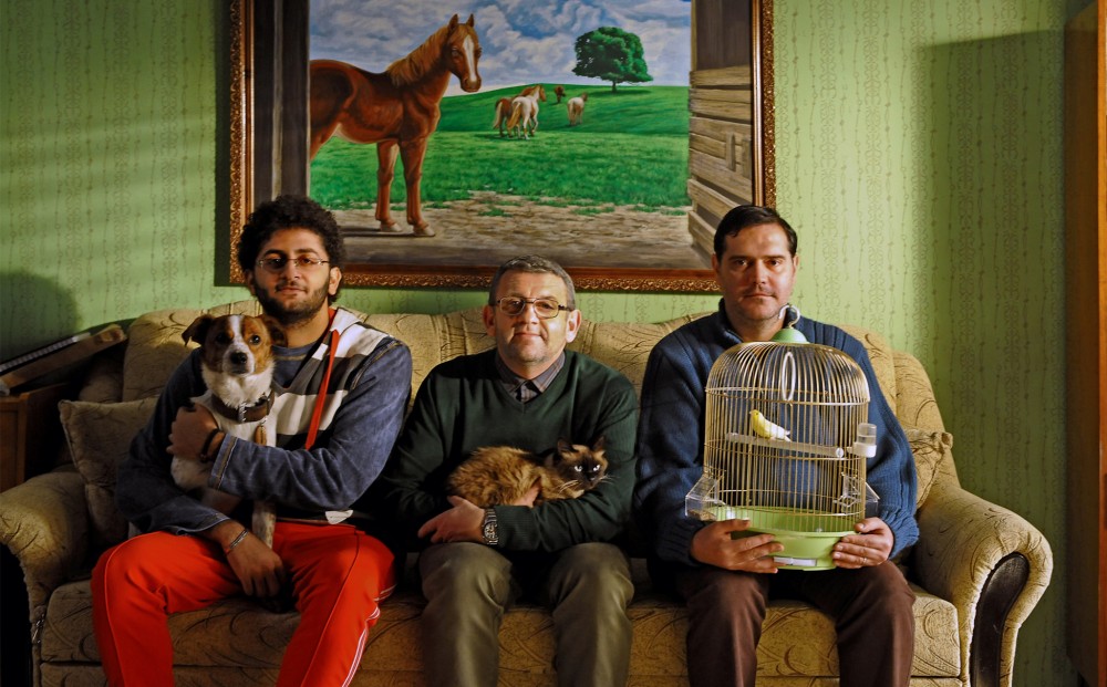 Three men sit next to each other on a couch; a painting of horses is on the wall behind them. One man holds a dog, one a cat, and the other a bird in a birdcage.