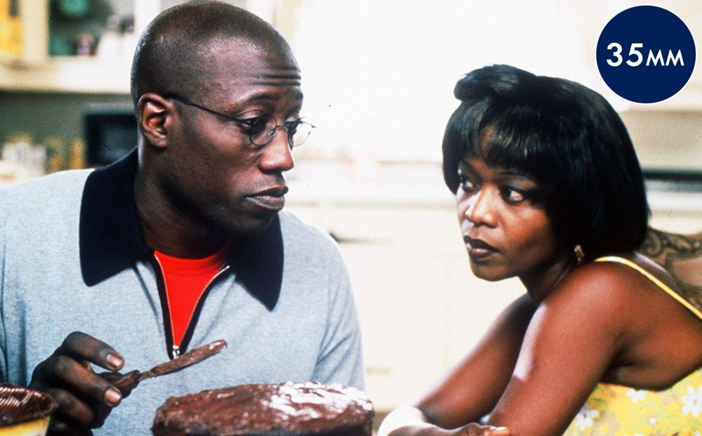 Actor Wesley Snipes frosts a chocolate cake and looks at Alfre Woodard, who sits next to him.