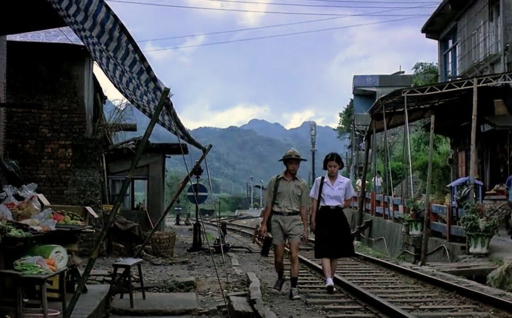 A young man and woman walk along train tracks that run through a marketplace.