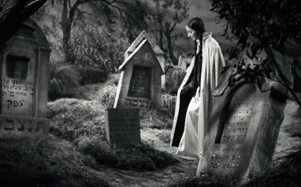 The pale woman with two long, dark braids stands in a cemetery in front of a headstone.