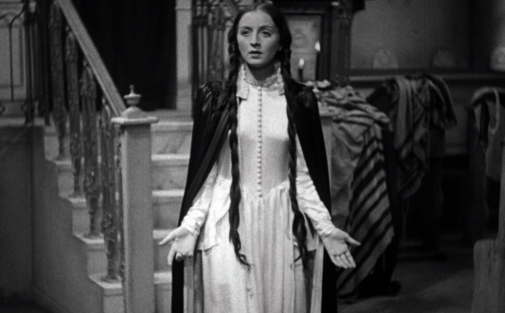 A pale woman with two long, dark braids stands in a gown with her arms by her sides, palms facing forward.