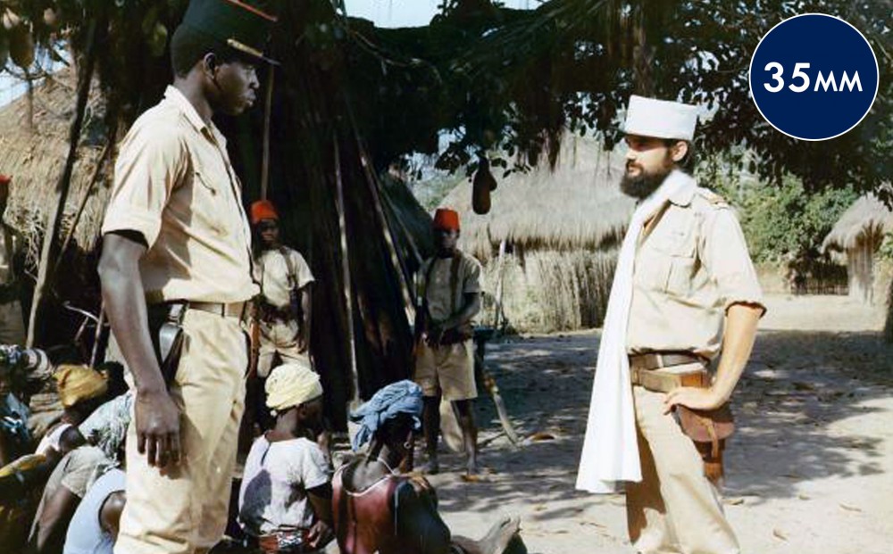 Two men in soldier's uniforms, one white and one black, seem to confront each other in a village.