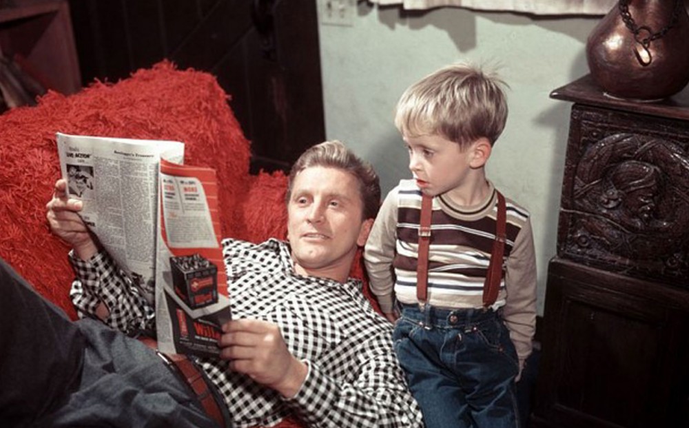 Kirk Douglas reads a newspaper while laying on a couch; a very young Michael Douglas sits next to him.