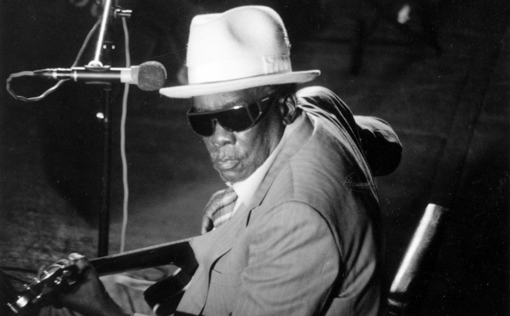 Musician Corey Harris sits in front of a microphone, holding a guitar and wearing a white fedora and sunglasses.