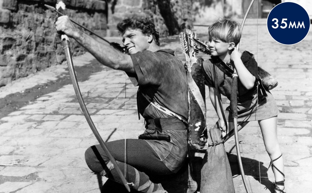 Actor Burt Lancaster kneels, aiming his bow and arrow upwards; a child who also has a bow and arrow stands next to him.