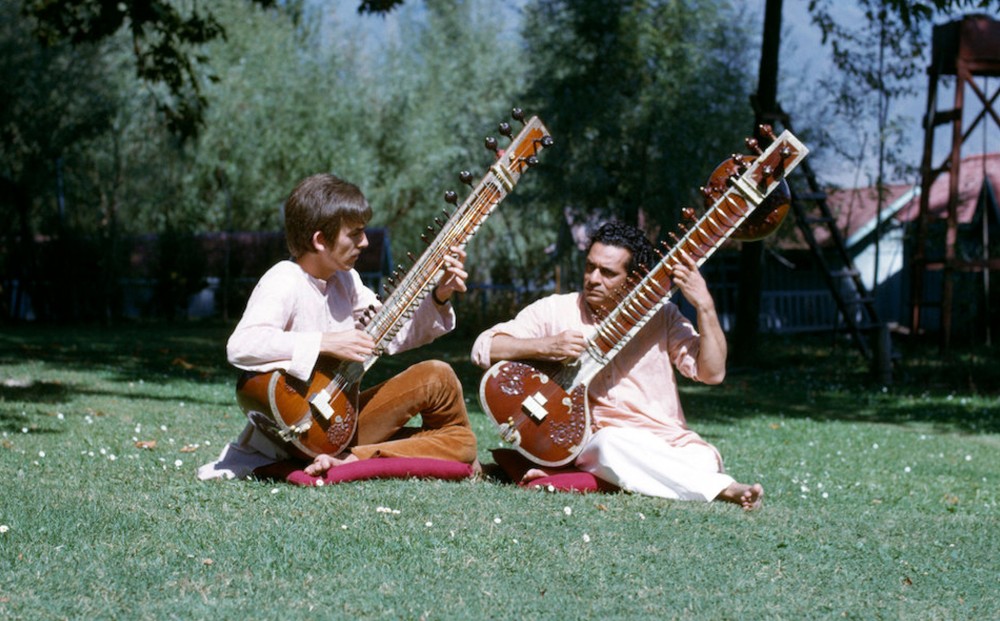 George Harrison and another man play sitars, seated on cushions on grass outside.