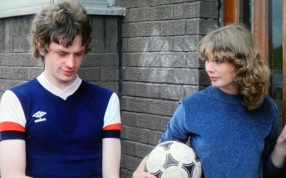 Actor Dee Hepburn holds a soccer ball and looks at John Gordon Sinclair, who looks down.