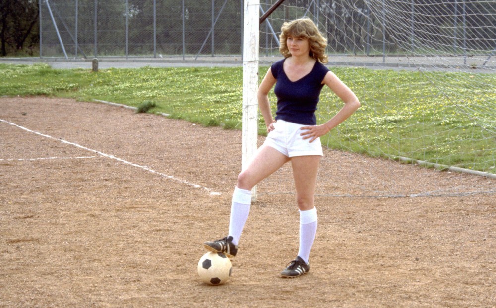 Actor Dee Hepburn stands with her hands on her hips and a soccer ball under her right foot.