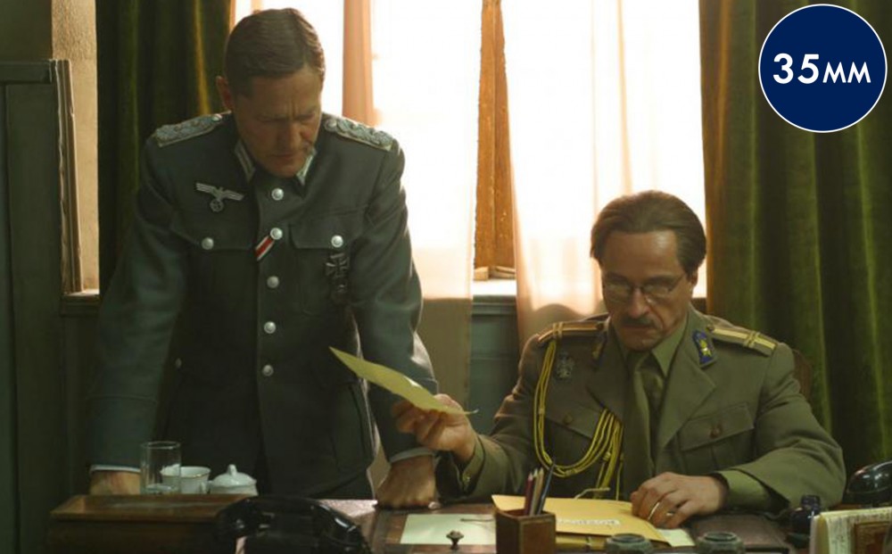 One man in an army uniform sits at a desk; another man in uniform next to him stands with his hands leaning on the desk.