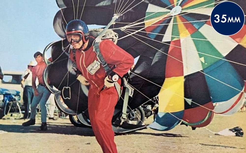 A man wears a skydiving helmet, goggles, and suit, with an expanded parachute on his back.