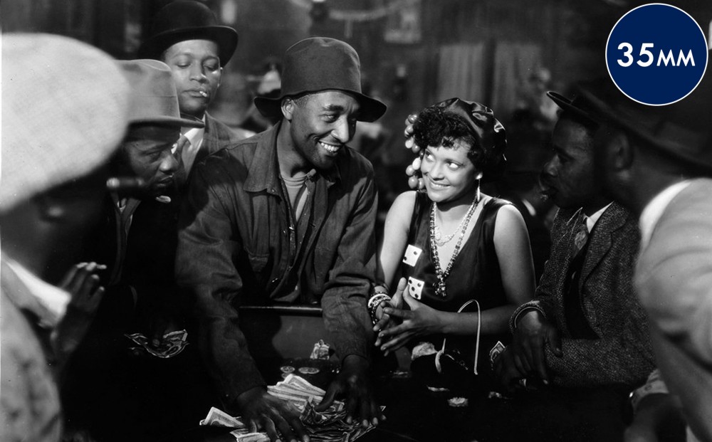 A woman stands around a gambling table with men, one of whom spreads cash around the table and smiles at her.