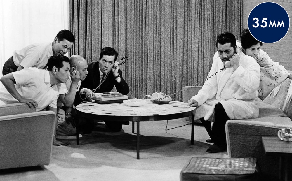 Four men sit on one side of a coffee table, and a man and a woman sit on the other, all listening to a conversation that one is having on the telephone.