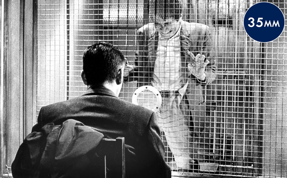 A man is seated in front of another man who grips the metal bars behind which he is imprisoned.
