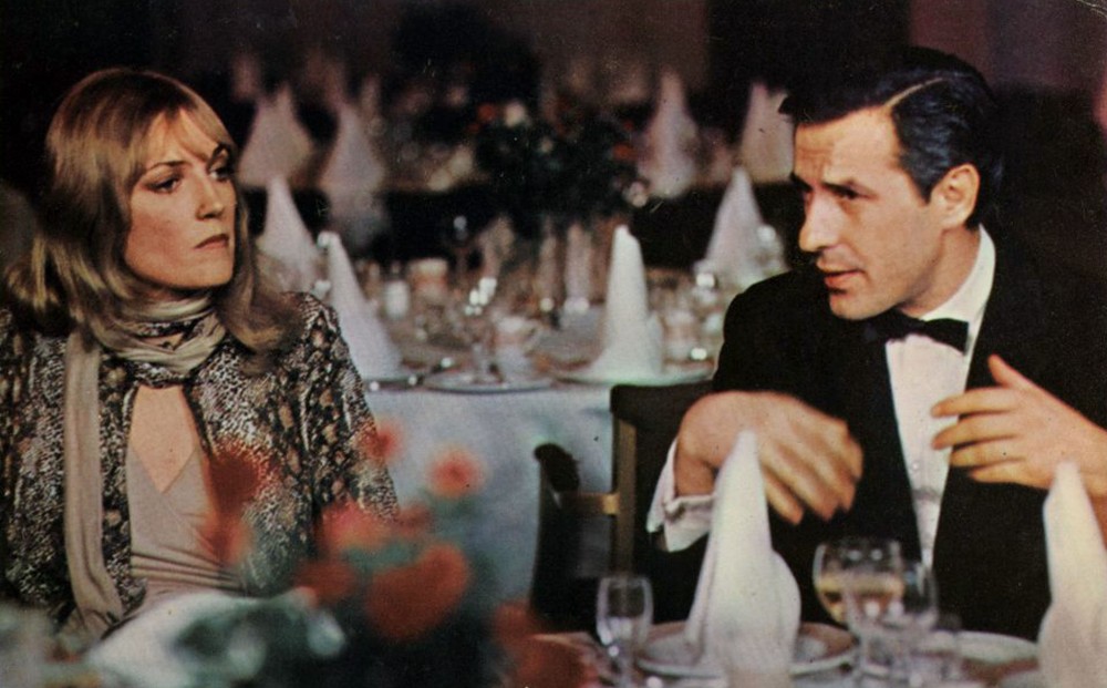 Actor John Cassavetes sits at a table with a woman in a fancy restaurant, wearing a suit.