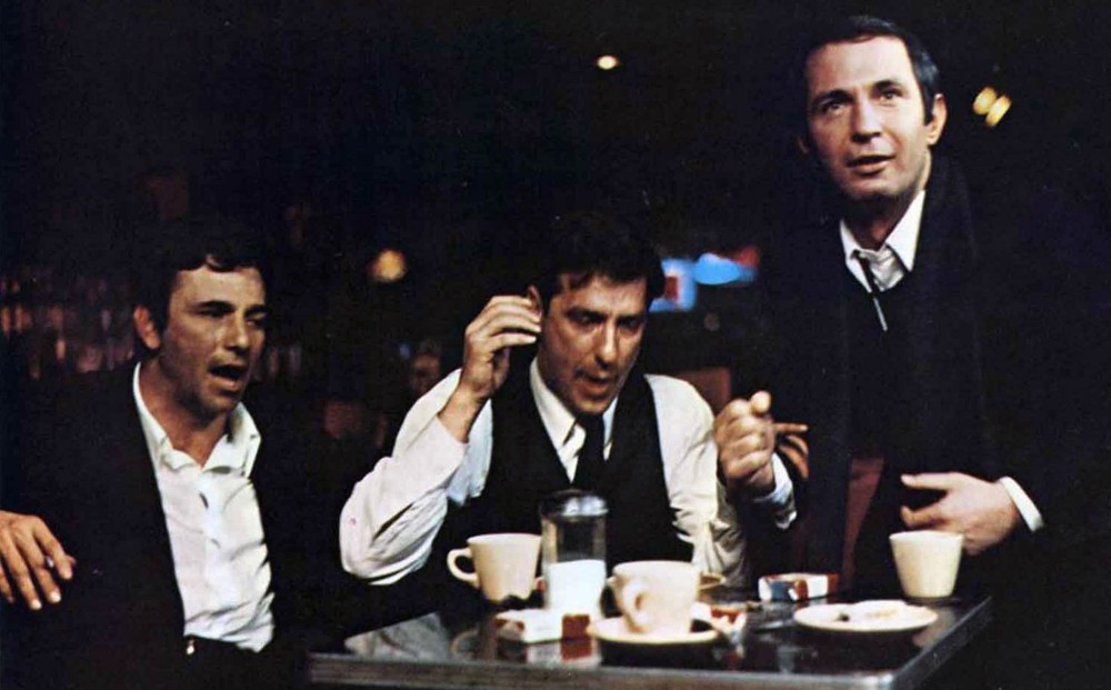 Actors John Cassavetes, Peter Falk, and Ben Gazzara sit together at a table, interacting animatedly.