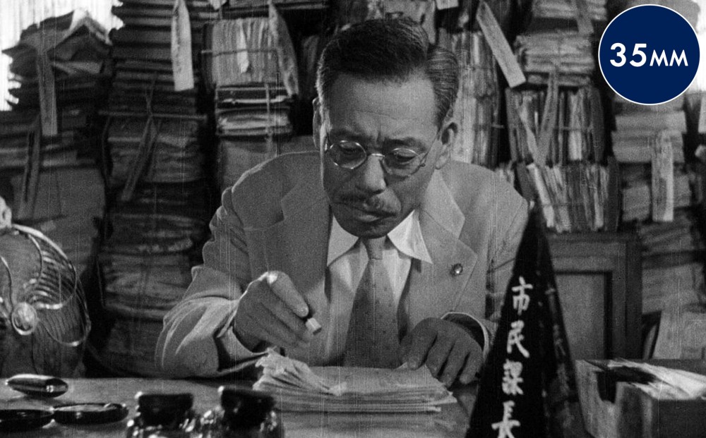 A man sits at a desk, surrounded by papers.
