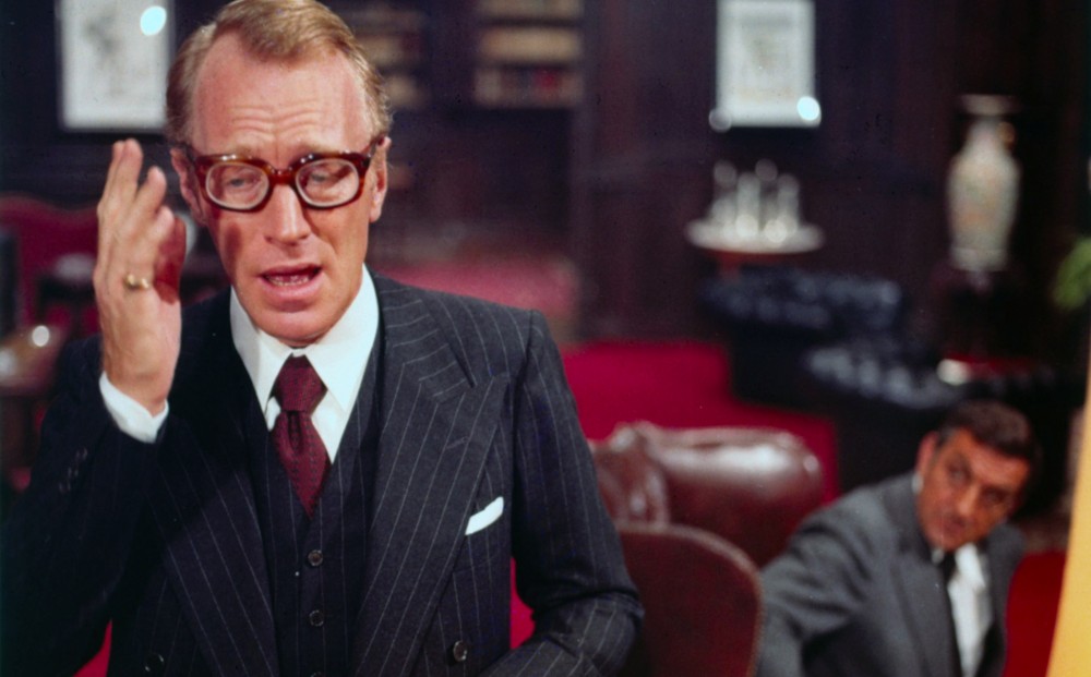 A man in a pin-striped three-piece suit speaks to a man who appears out-of-focus in the background.