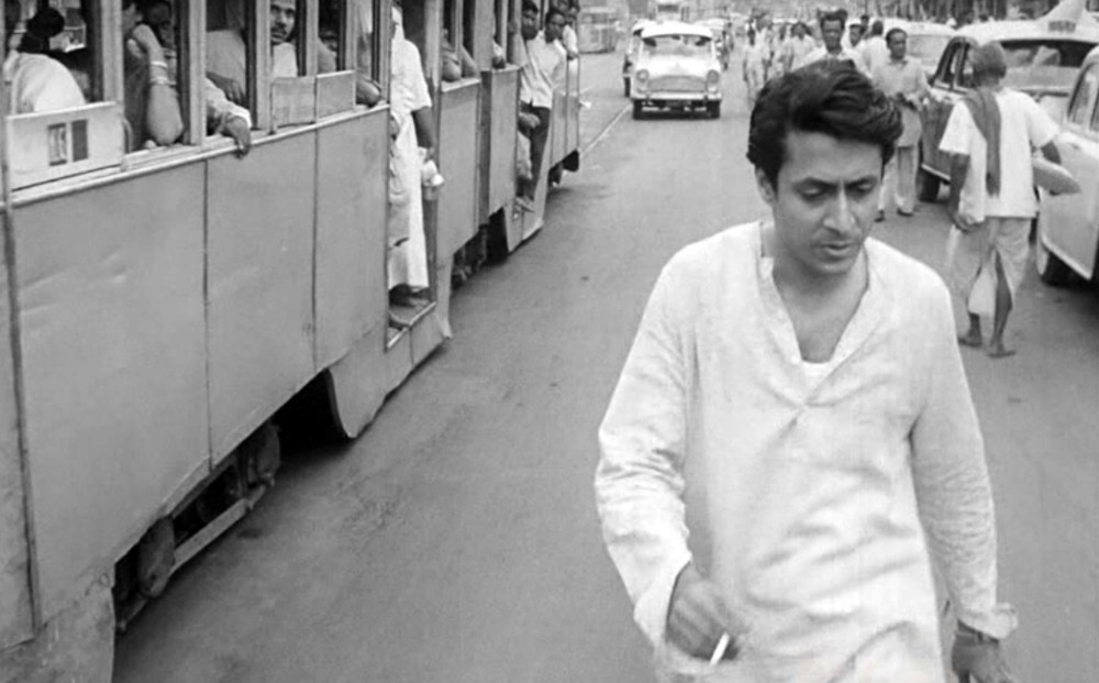 Ranjit Mallick walks alongside a bus in the road, holding a cigarette.