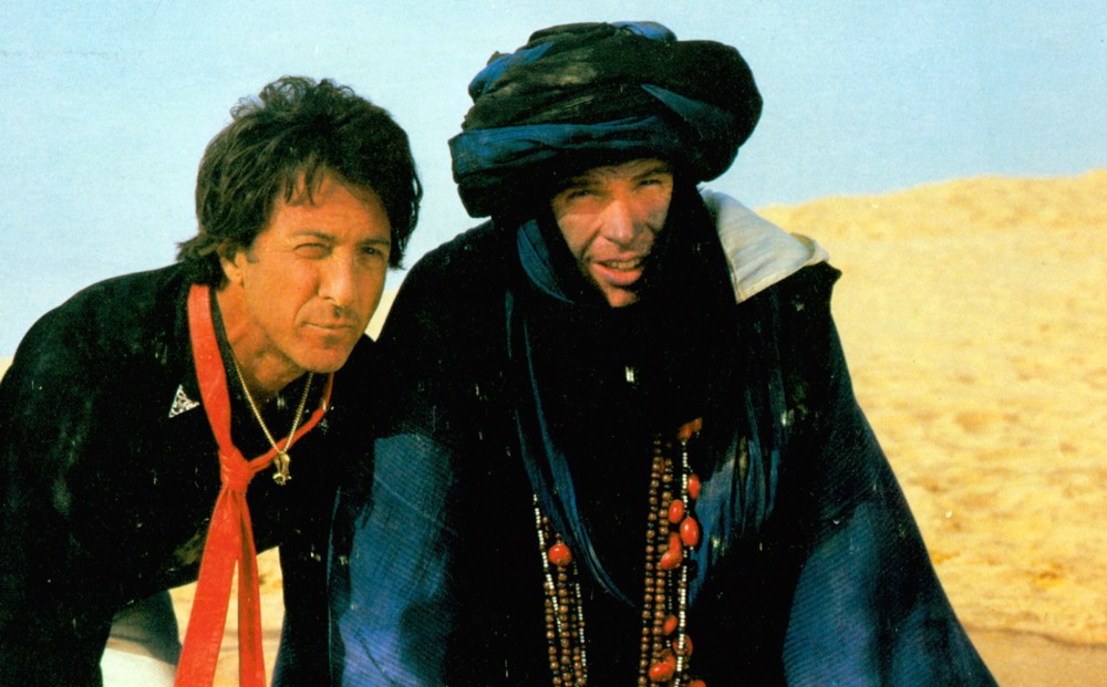Actors Dustin Hoffman and Warren Beatty crouch on the sand in the desert, the latter wearing a robe and turban.