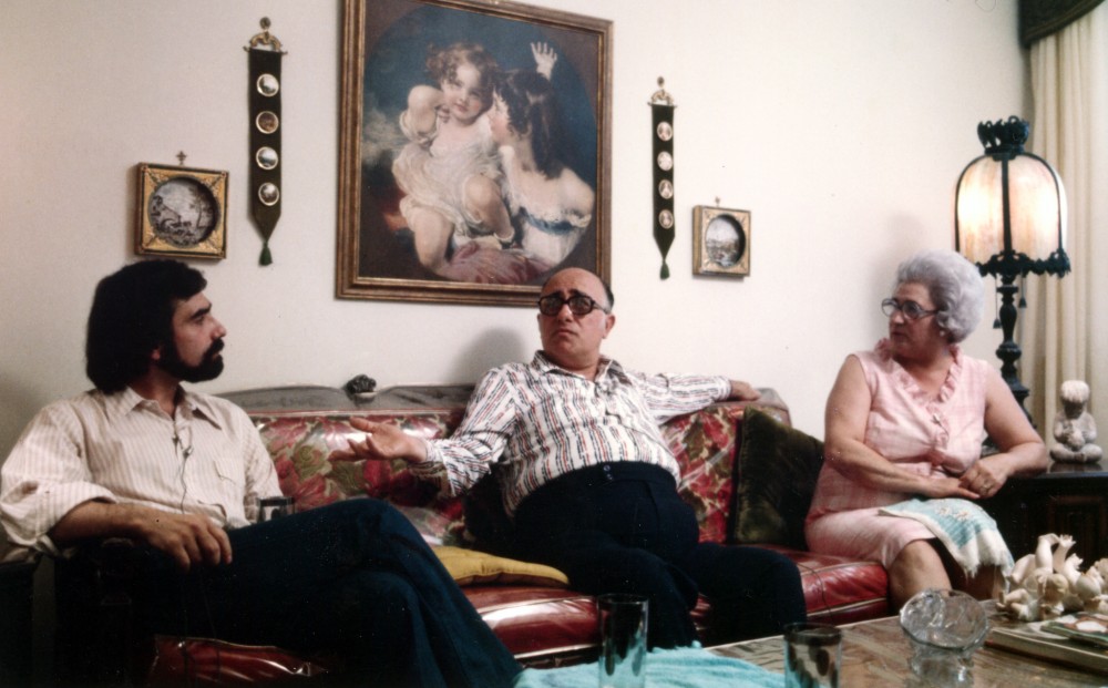 A middle-aged man, an older man, and an older woman sit in a living room on a couch with a plastic cover.