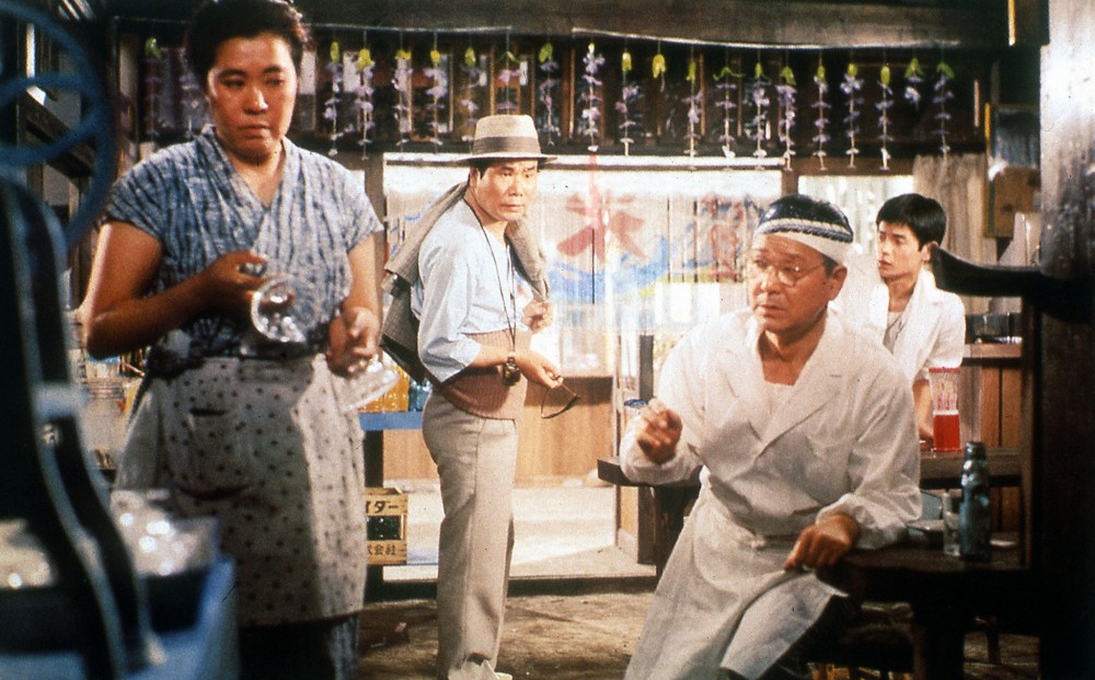 Actor Kiyoshi Atsumi stands inside of a business establishment with two men and a woman who all appear to work there.