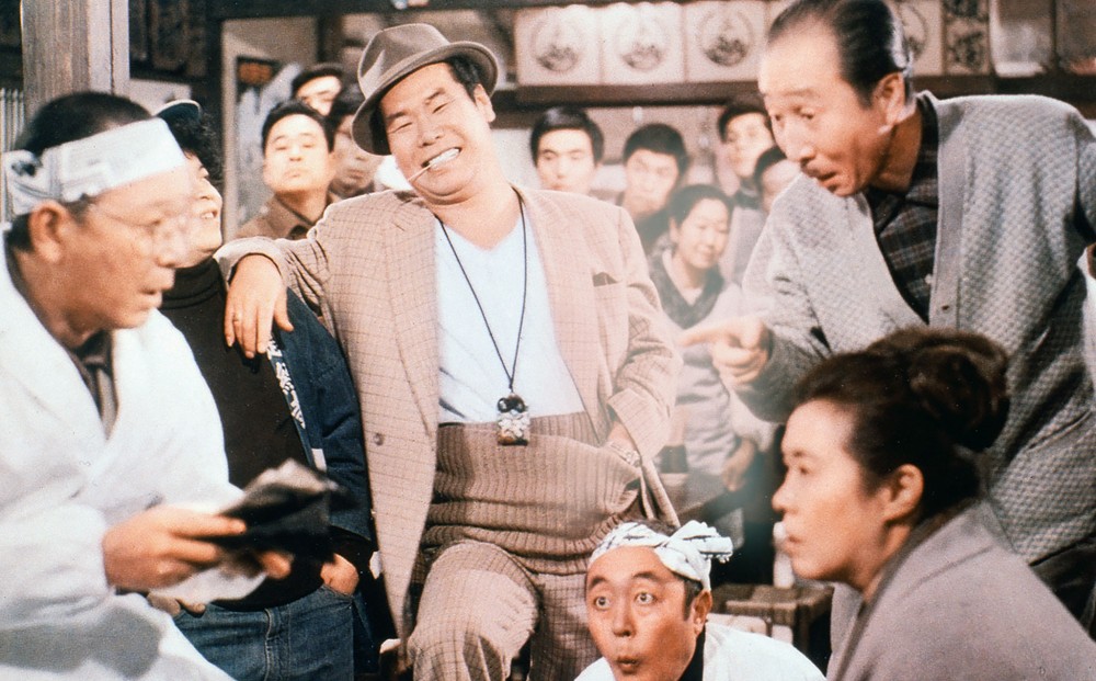 Actor Kiyoshi Atsumi smiles with a toothpick in his mouth, in a crowded room.