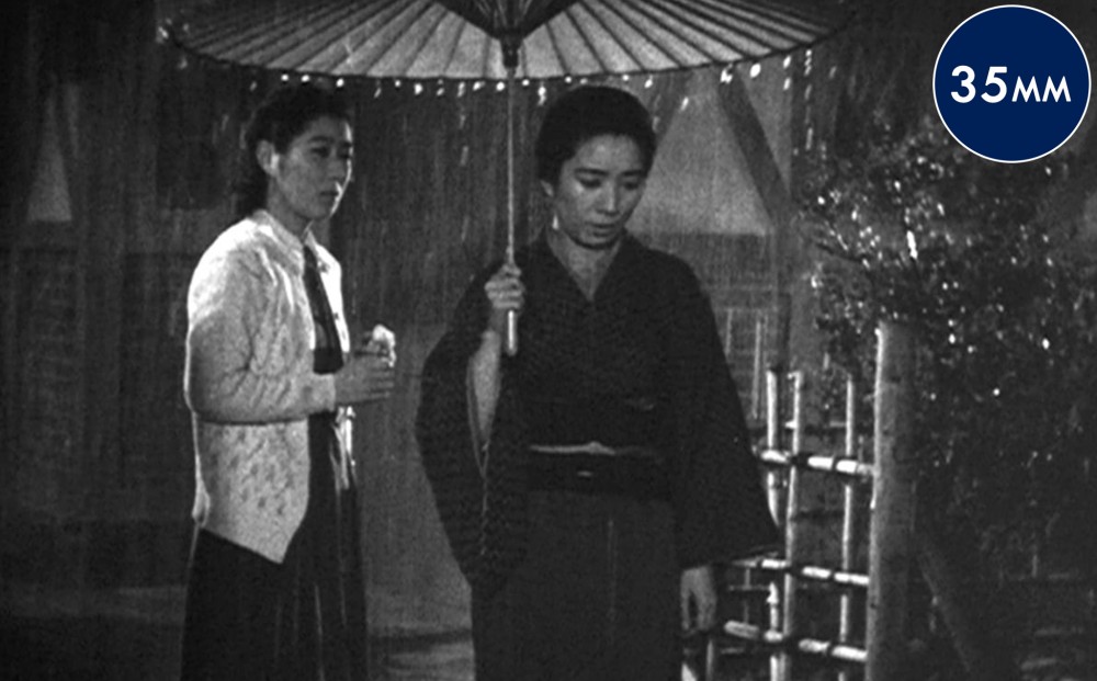 Two women stand outside in the rain; one is under an umbrella.