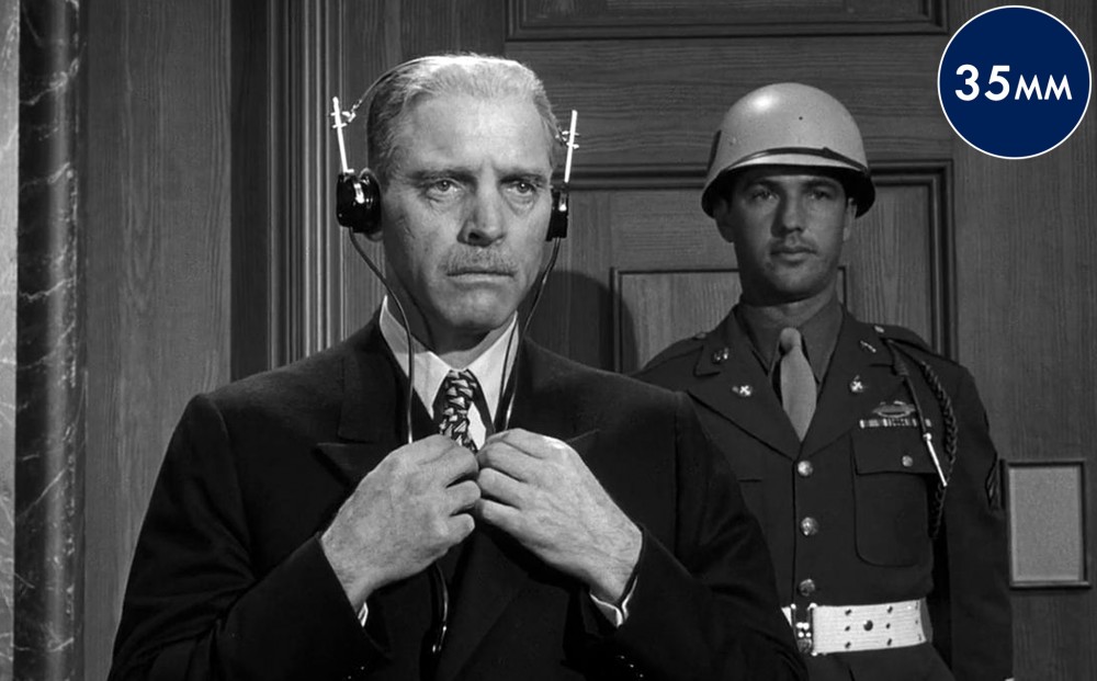 Actor Burt Lancaster stands in a courtroom, a soldier standing guard behind him.
