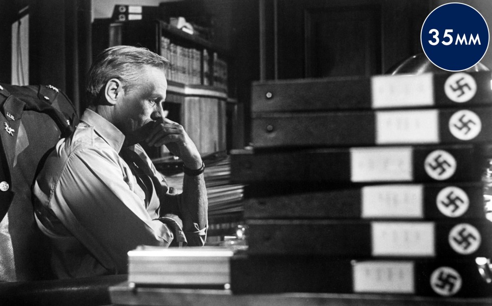 A man sits in an office, surrounded by binders with swastika labels.
